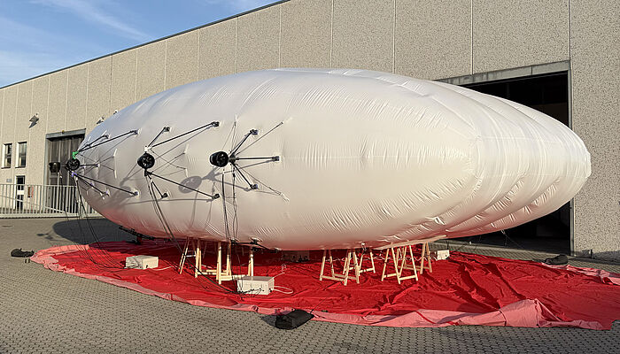 一艘来自 FloFleet 公司的白色飞艇停放在一个工业建筑前的红色防水布上。飞艇外部安装了多个由细长支架固定的螺旋桨装置。