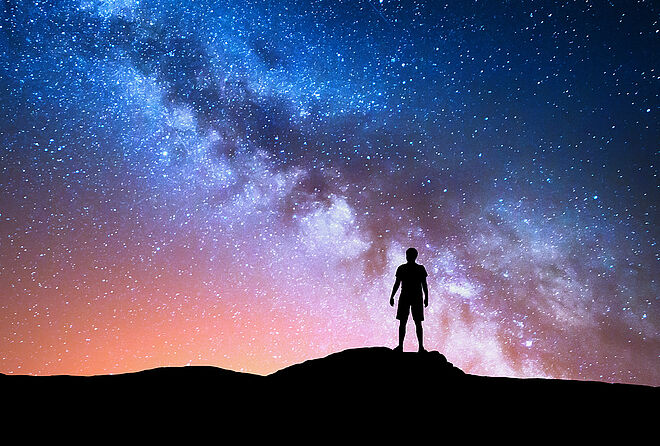 一个人站在高地上的剪影，头顶是晴朗的星空，清晰可见的银河横贯天际。该图展示了借助现代空间望远镜对宇宙进行观测的场景。
