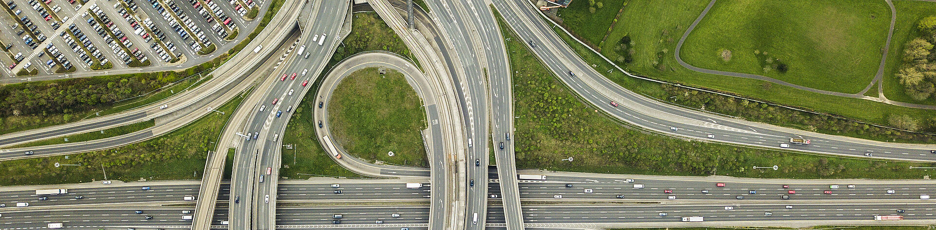 一张高速公路立交桥的俯视图，展示了多条车道、上下匝道。画面中还可见周围的停车场、建筑物和绿地，整体场景呈现出大规模基础设施的印象。