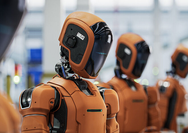 A row of orange humanoid robots with dark visor heads is shown in a modern manufacturing environment. The scene conveys the impression of an industrial production line in which humanoid robots are actively involved in automated processes.